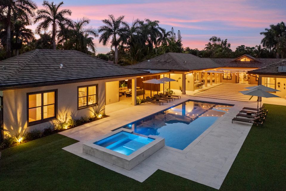 An illuminated home, garden and swimming pool surrounded by palms at dusk